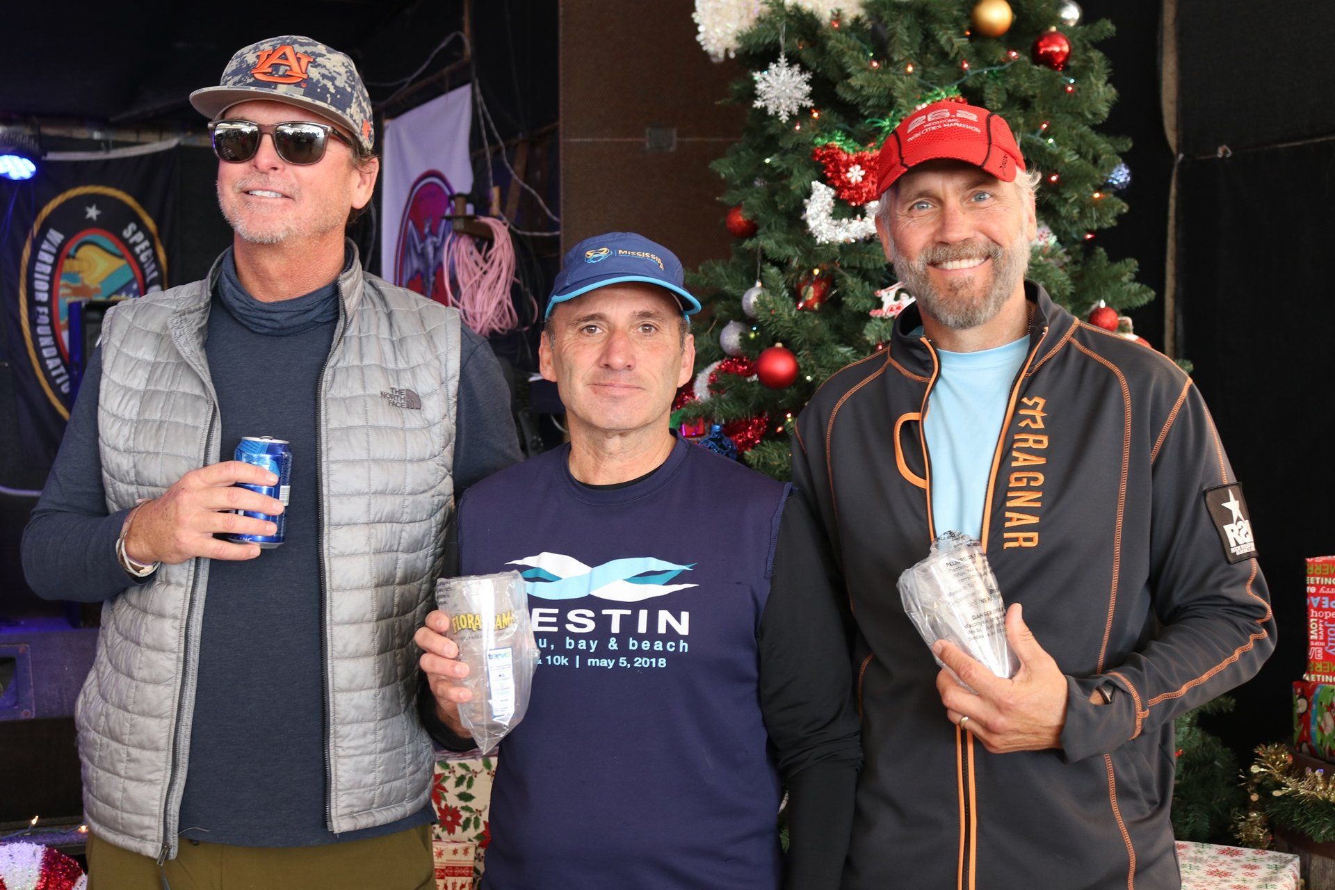 Three men holding drinks and awards pose together in front of a decorated Christmas tree.