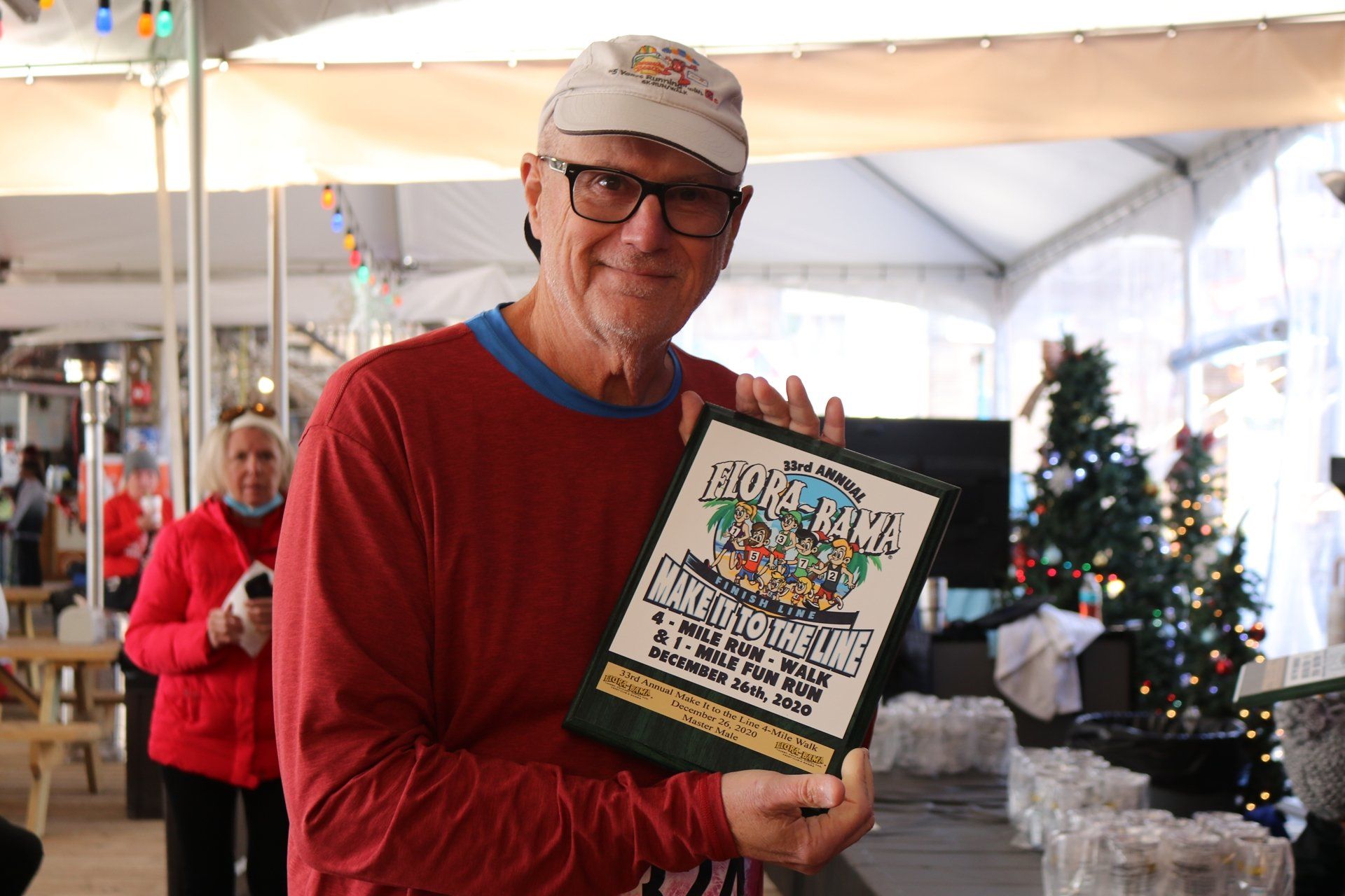 A person in a red shirt and baseball cap smiles while holding a commemorative event plaque under a white tent.