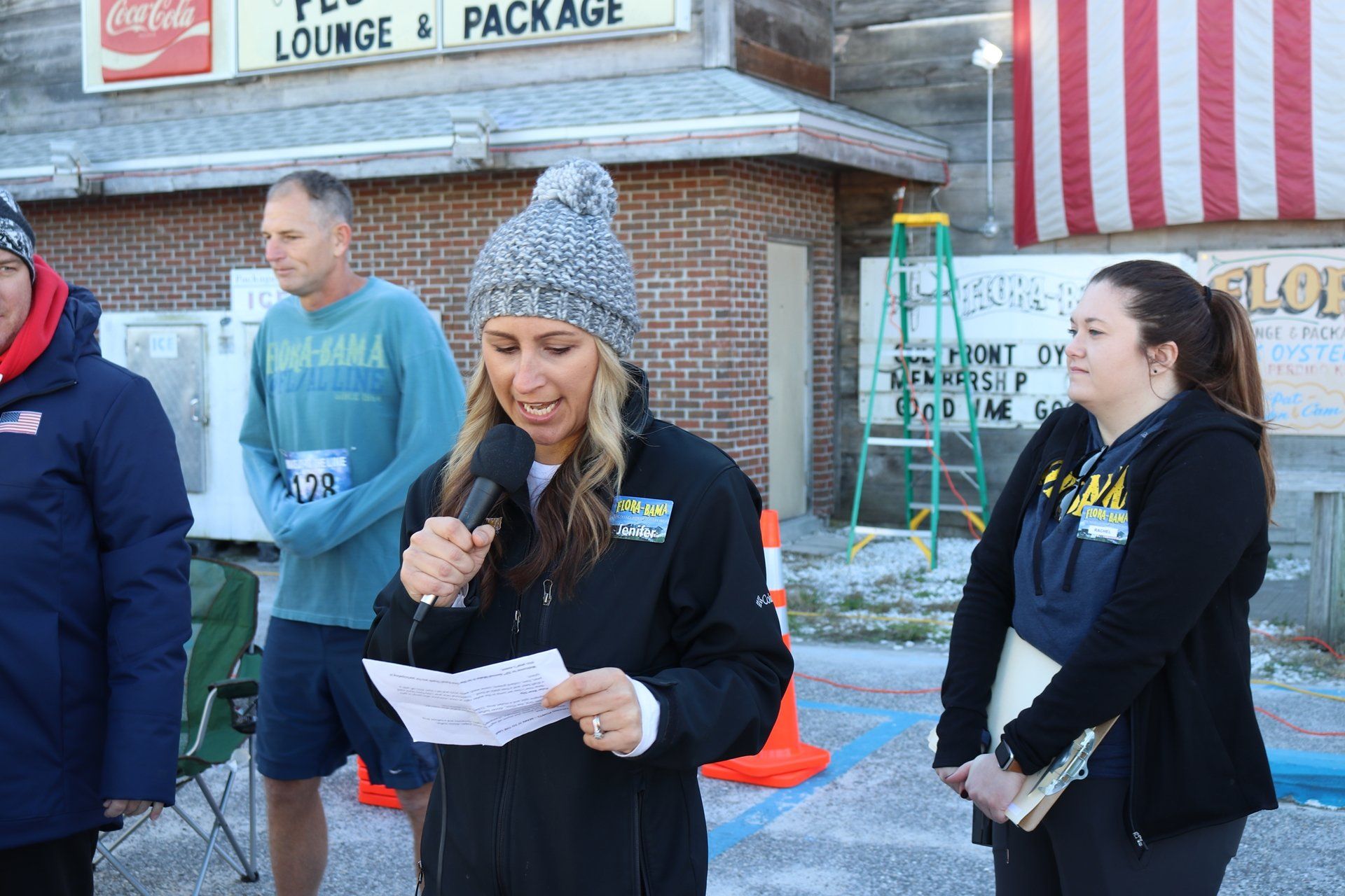 A woman in a grey beanie speaks into a microphone at an outdoor event, with two people standing behind her.