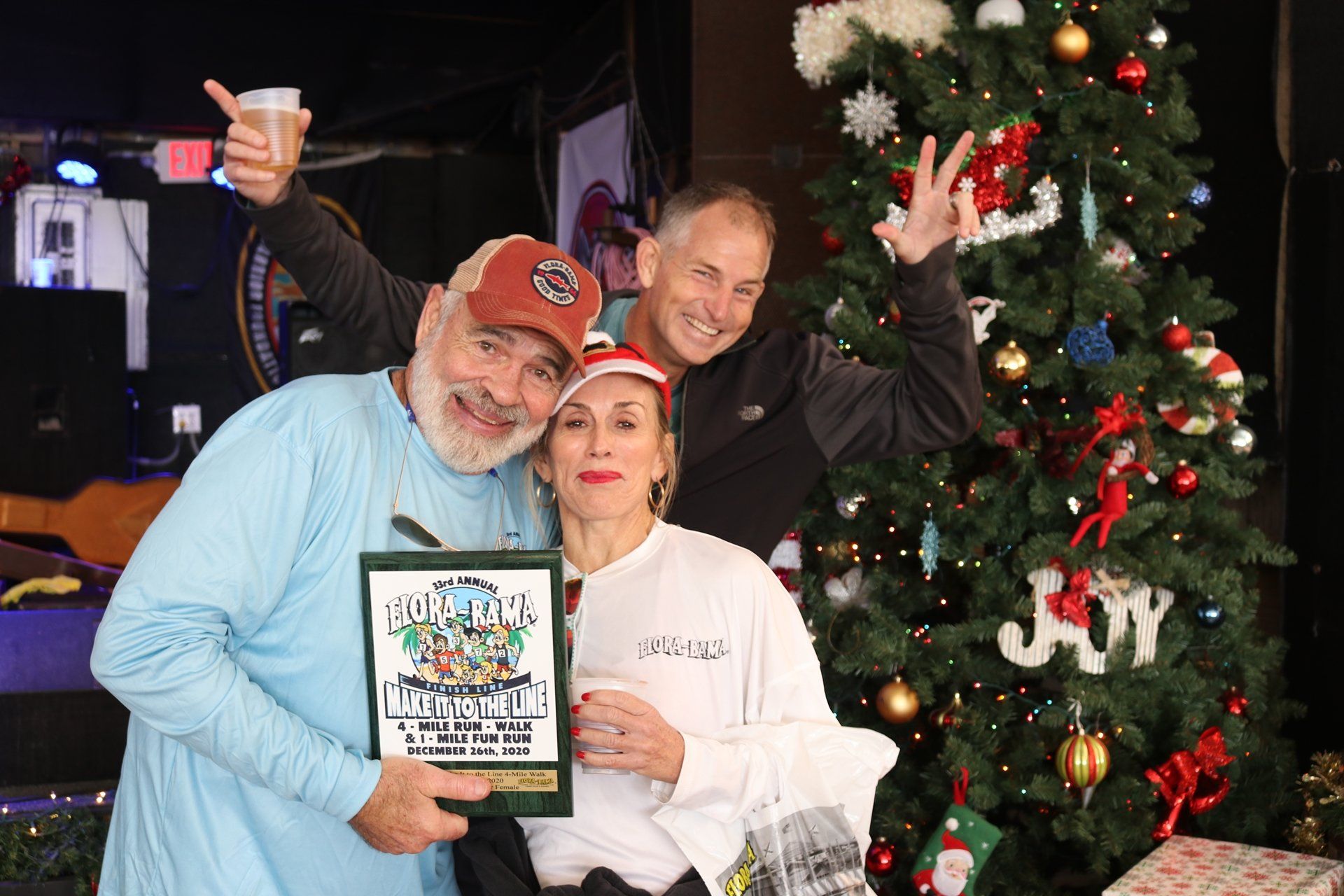 Three people pose happily in front of a Christmas tree, with one holding a framed award or sign.
