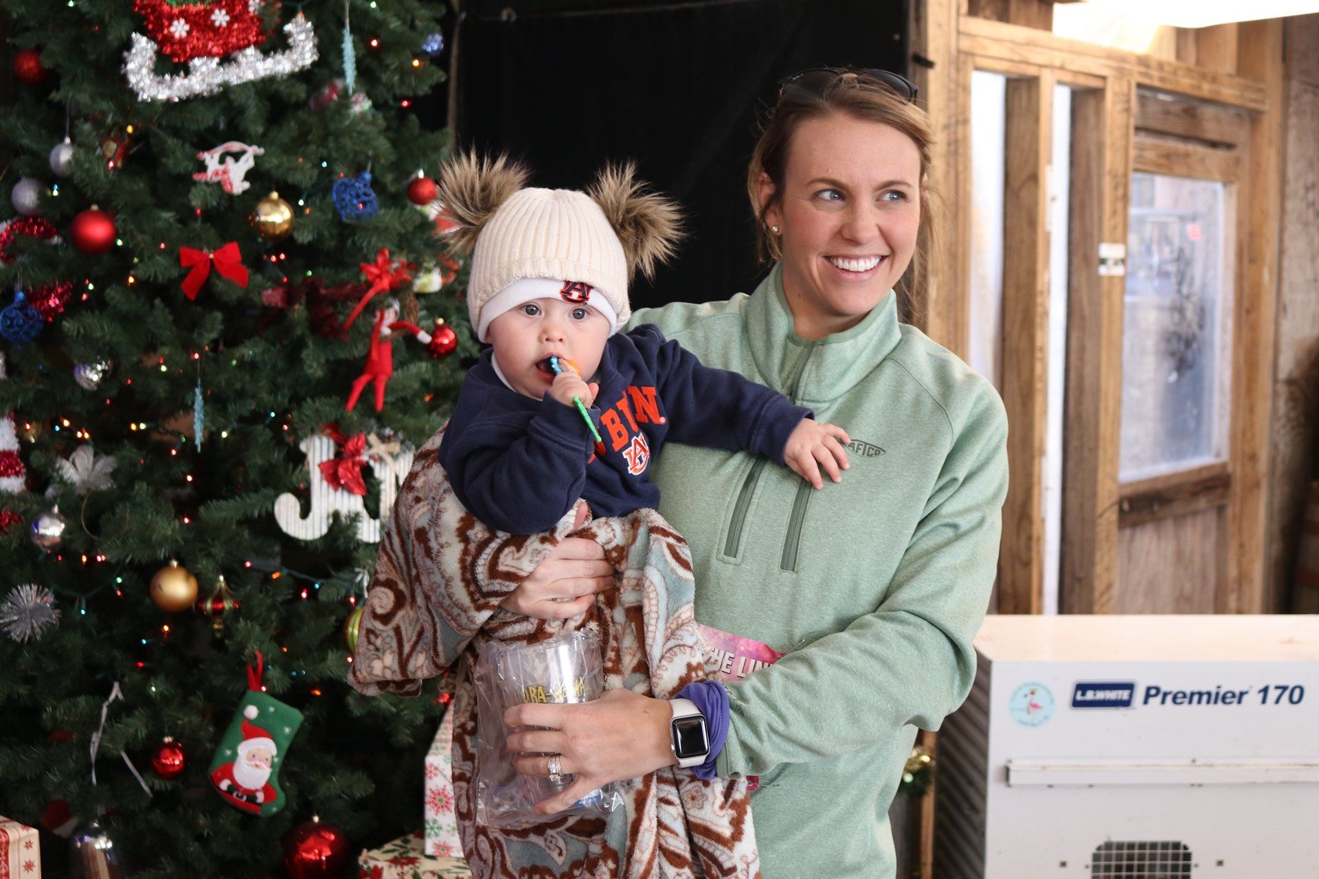 A person smiles while holding a baby in a patterned blanket near a decorated Christmas tree.