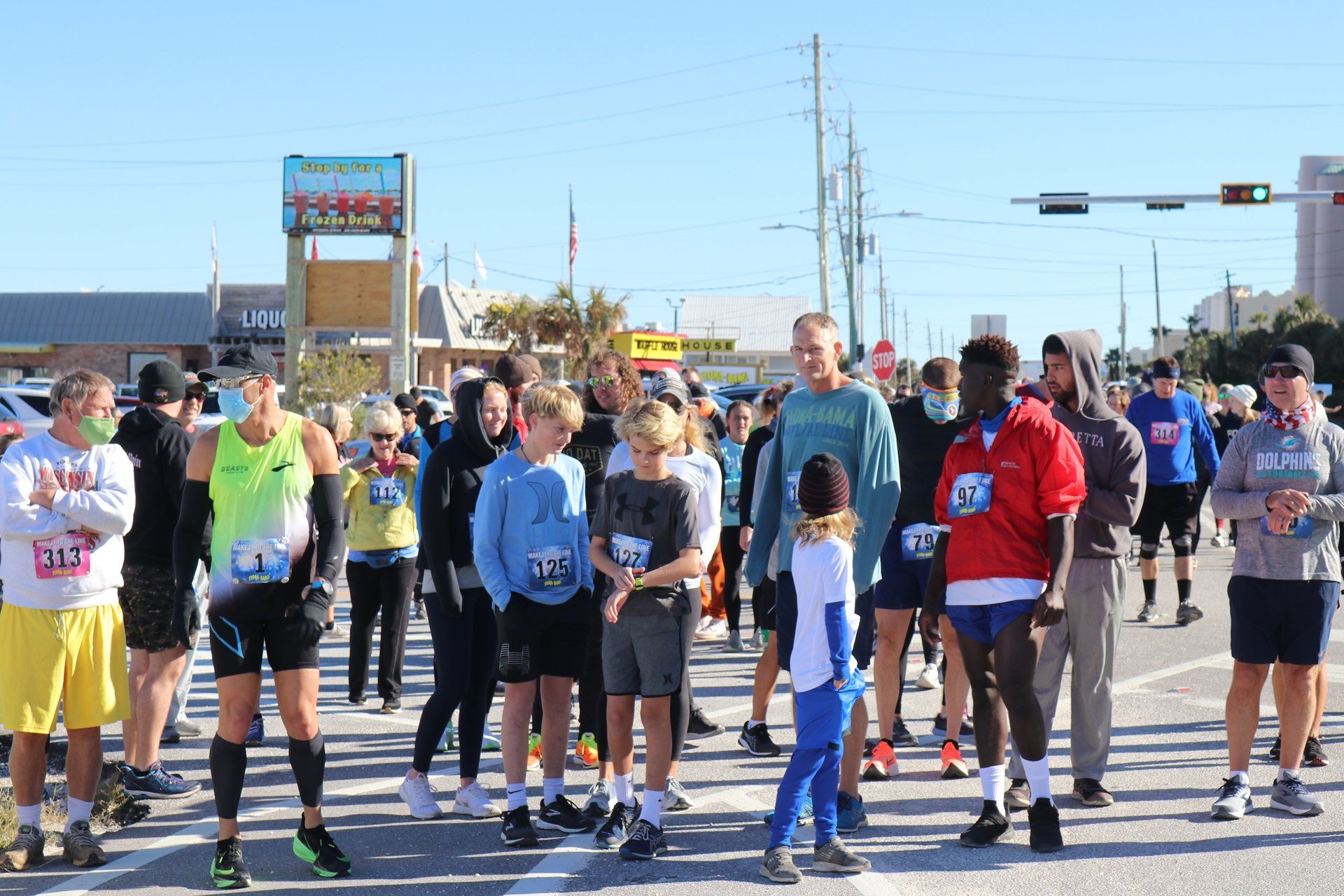A group of runners stands on a paved road during a sunny daytime race event, preparing to start or waiting in a crowd.