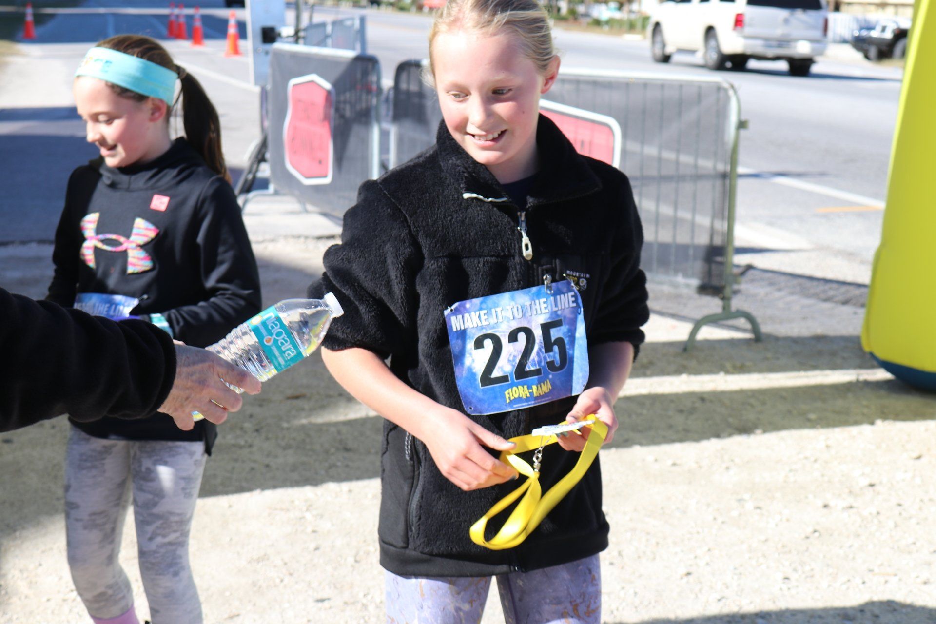 Two individuals wearing race bibs outdoors; one holds a water bottle while the other looks at a yellow medal.