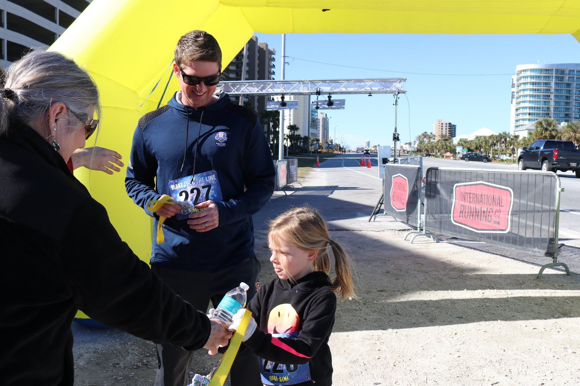 A volunteer hands a yellow finisher medal to a young runner under a bright yellow archway at a race finish line.