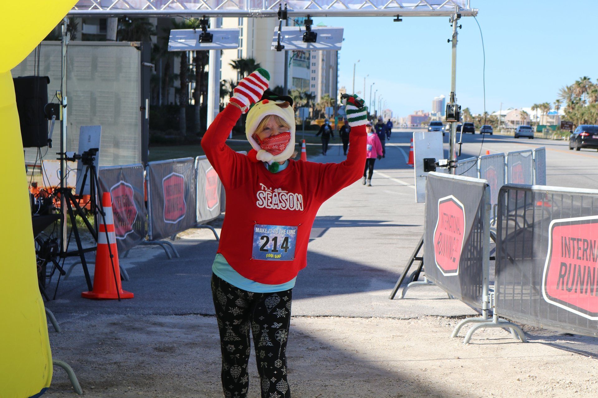 A runner wearing a red long-sleeve shirt and mask with a race bib celebrates at a finish line with arms raised.