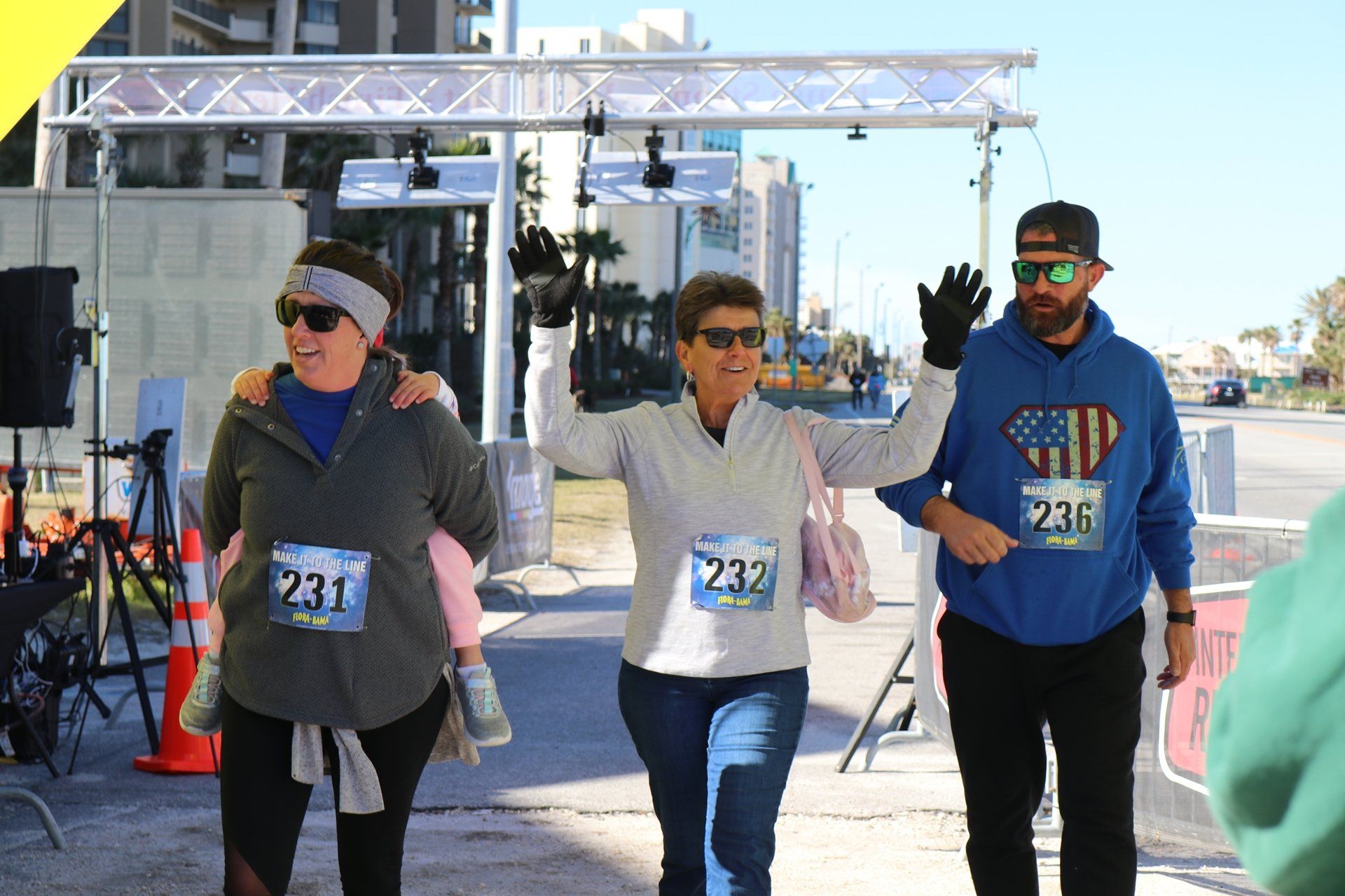 Three people wearing race bibs walk past a finish line arch, with the middle person waving and a child riding piggyback.