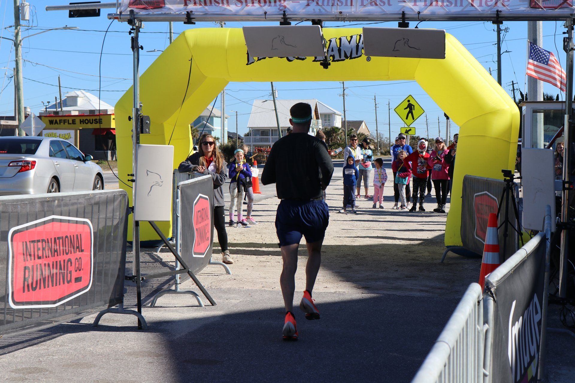 A runner crosses the finish line under a yellow inflatable arch at an outdoor race event with spectators in the background.