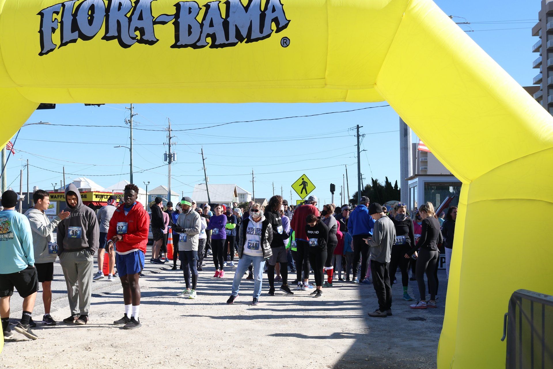 A large, yellow inflatable Flora-Bama arch stands at the start of an outdoor running race with many participants nearby.