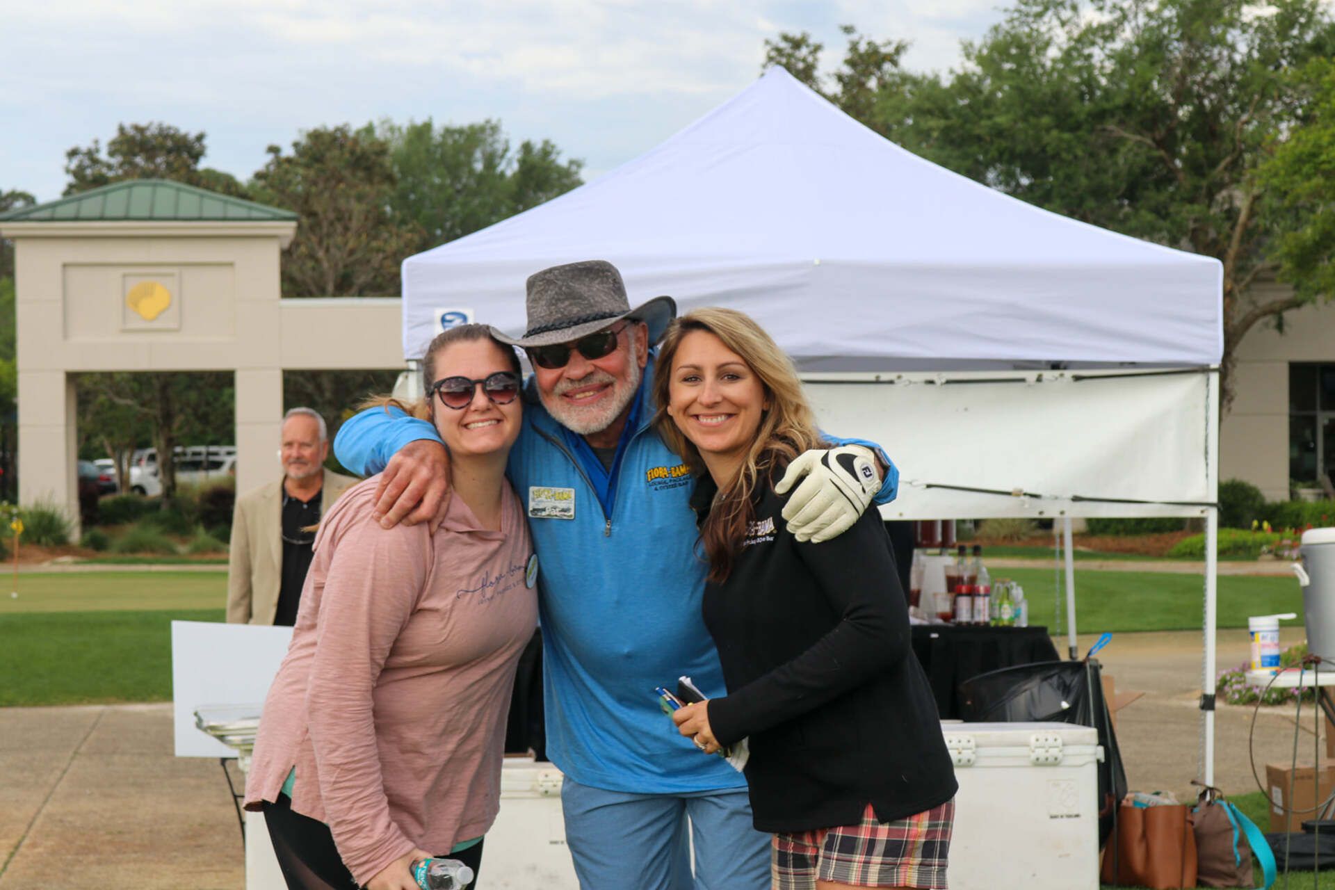 Three people pose for a photo in front of a white tent on a sunny, outdoor event lawn.