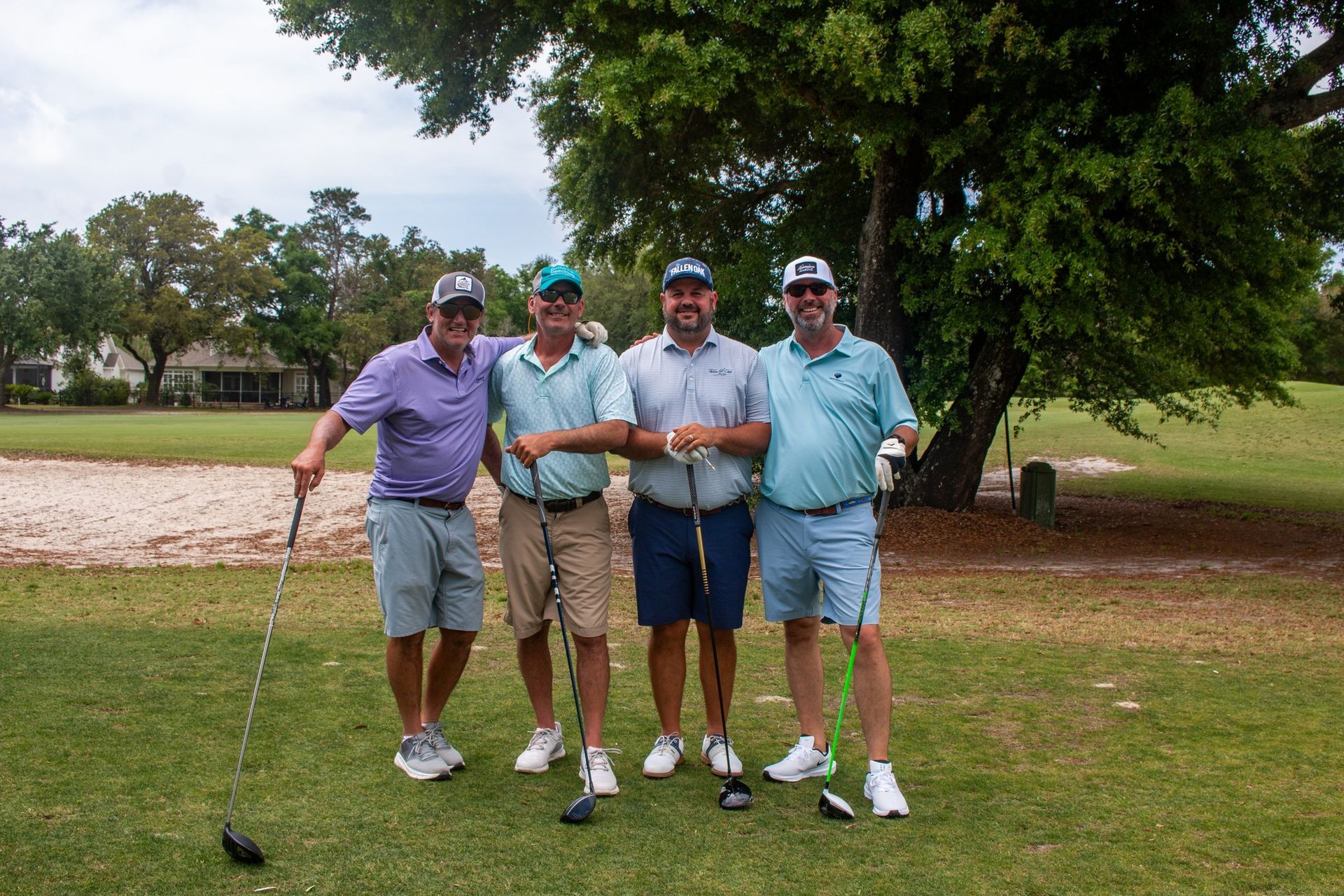 Four people wearing golf attire pose with golf clubs on a grassy course in front of a large tree.