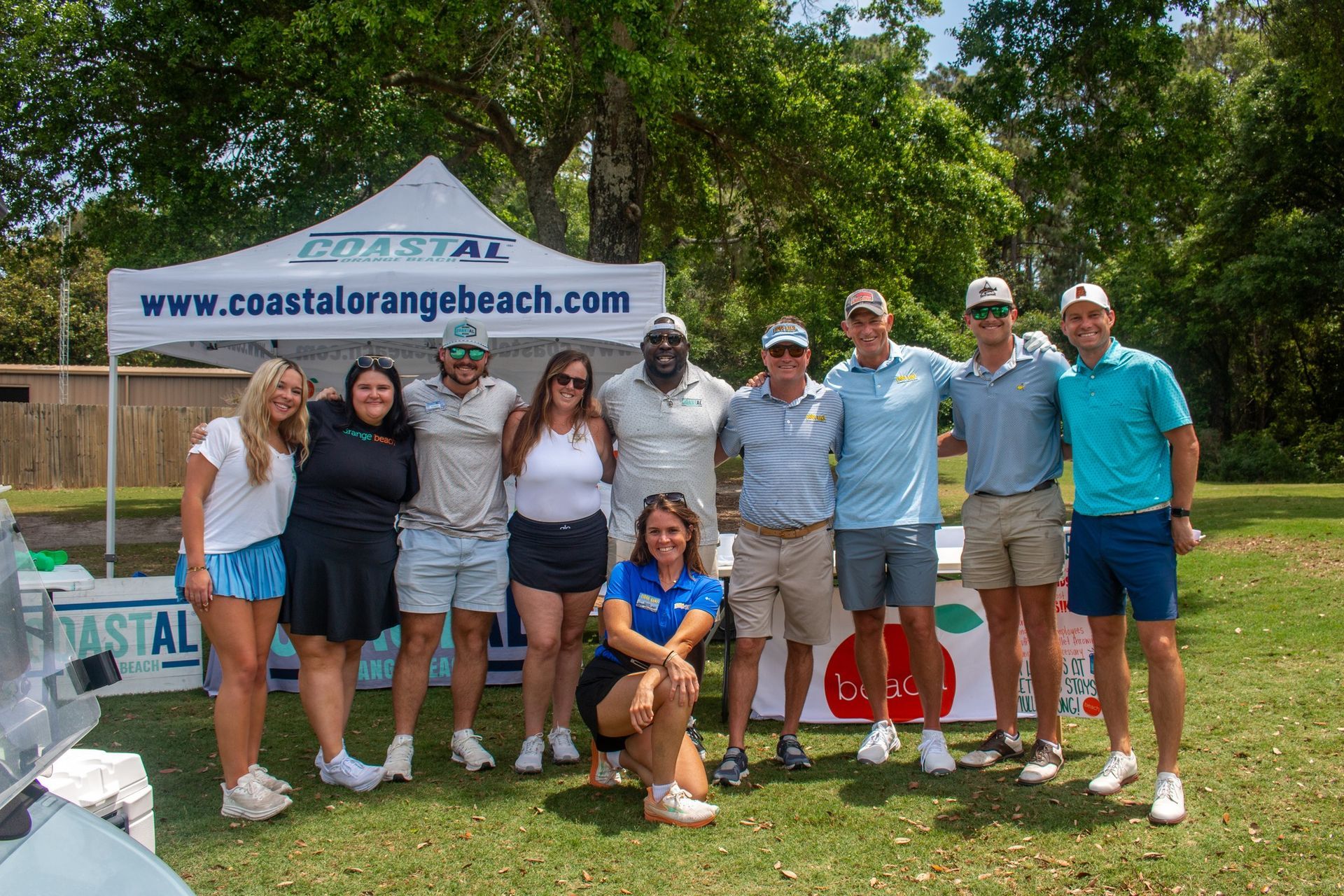 A group of ten people stand smiling under a Coastal Orange Beach event tent in a grassy outdoor setting.