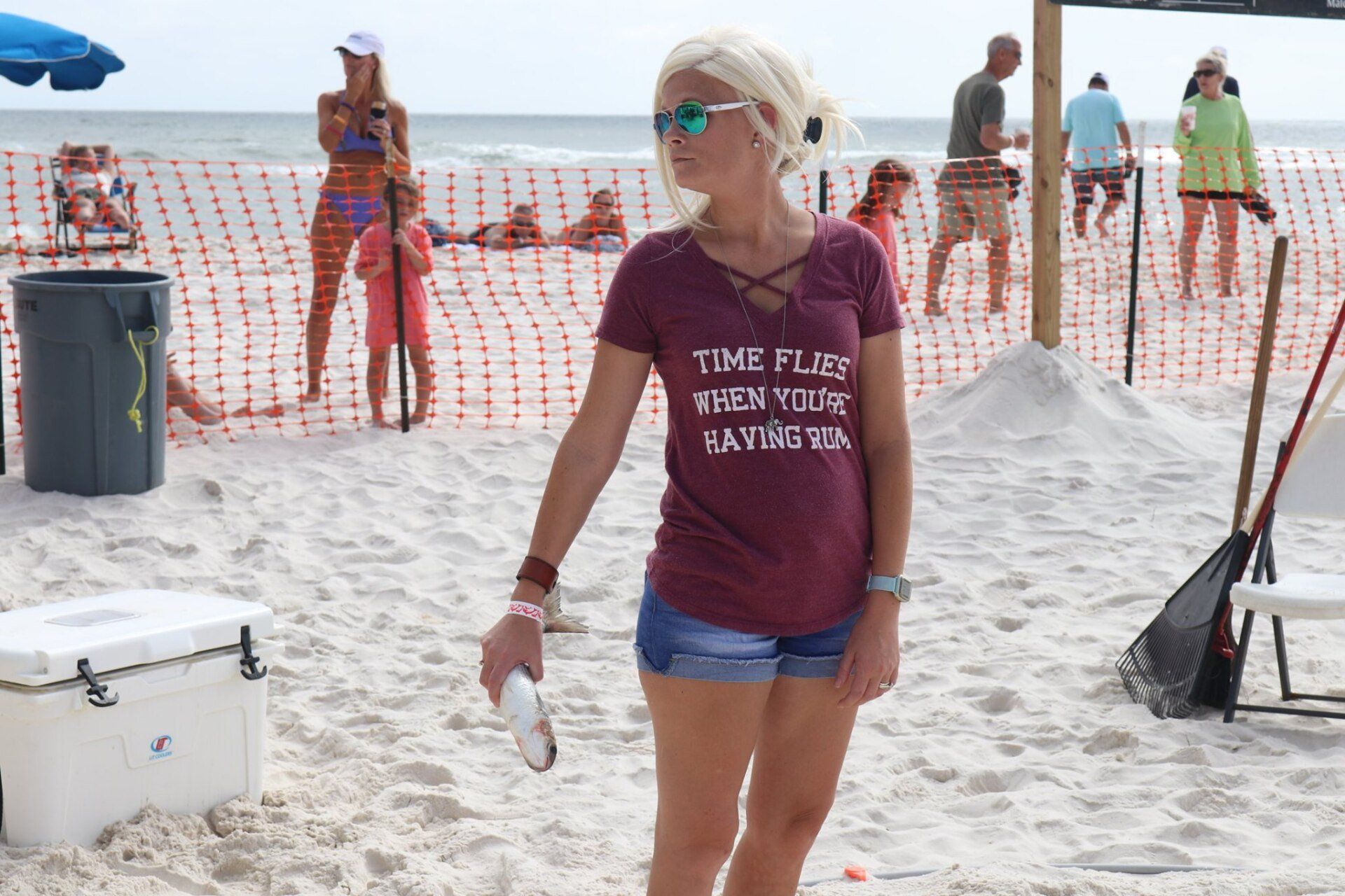 A person wearing a maroon t-shirt and denim shorts holds a drink on a sandy beach near a crowd and a cooler.