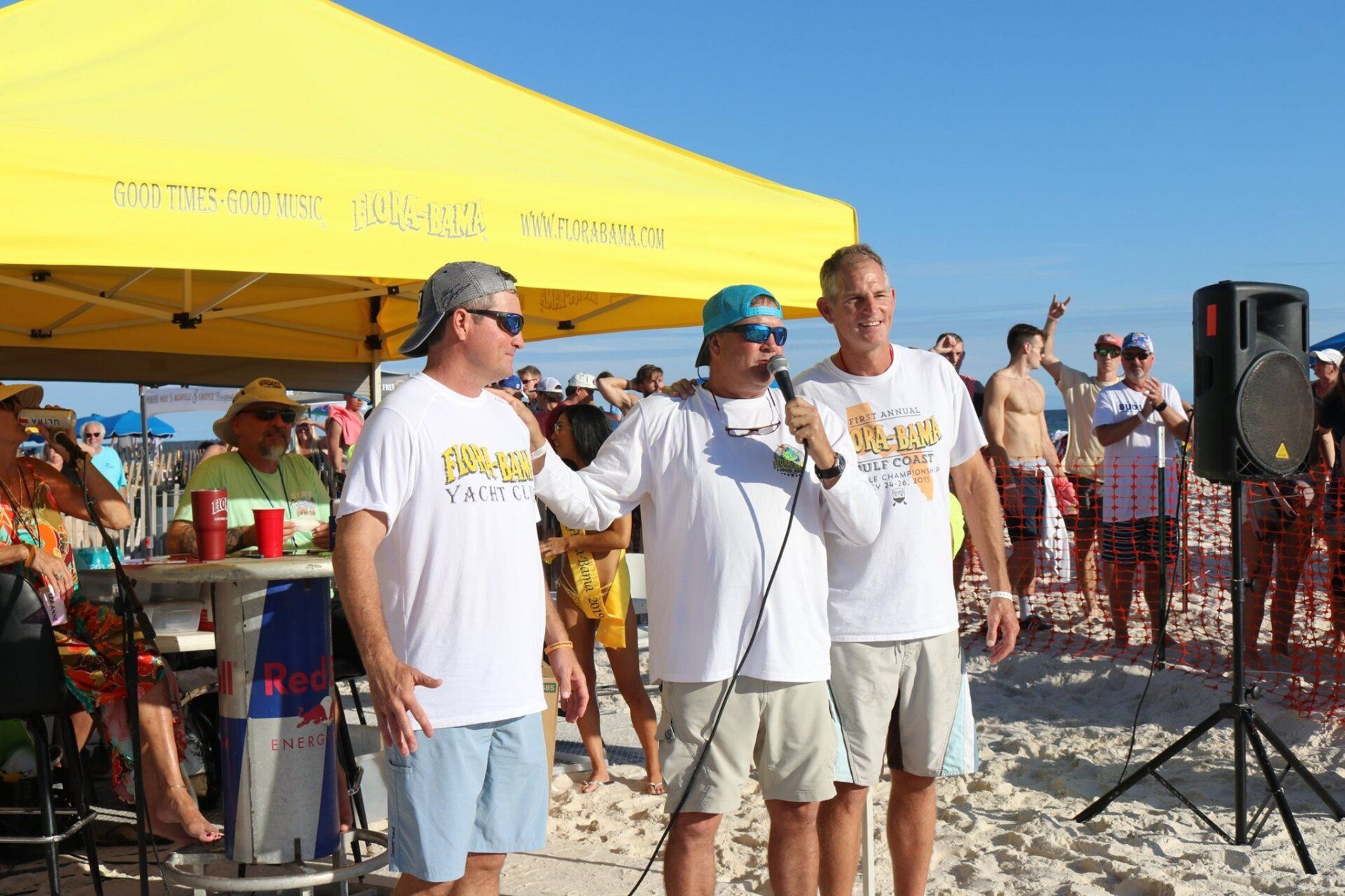 Three people stand under a yellow tent on a sunny beach, one speaking into a microphone while others look on.