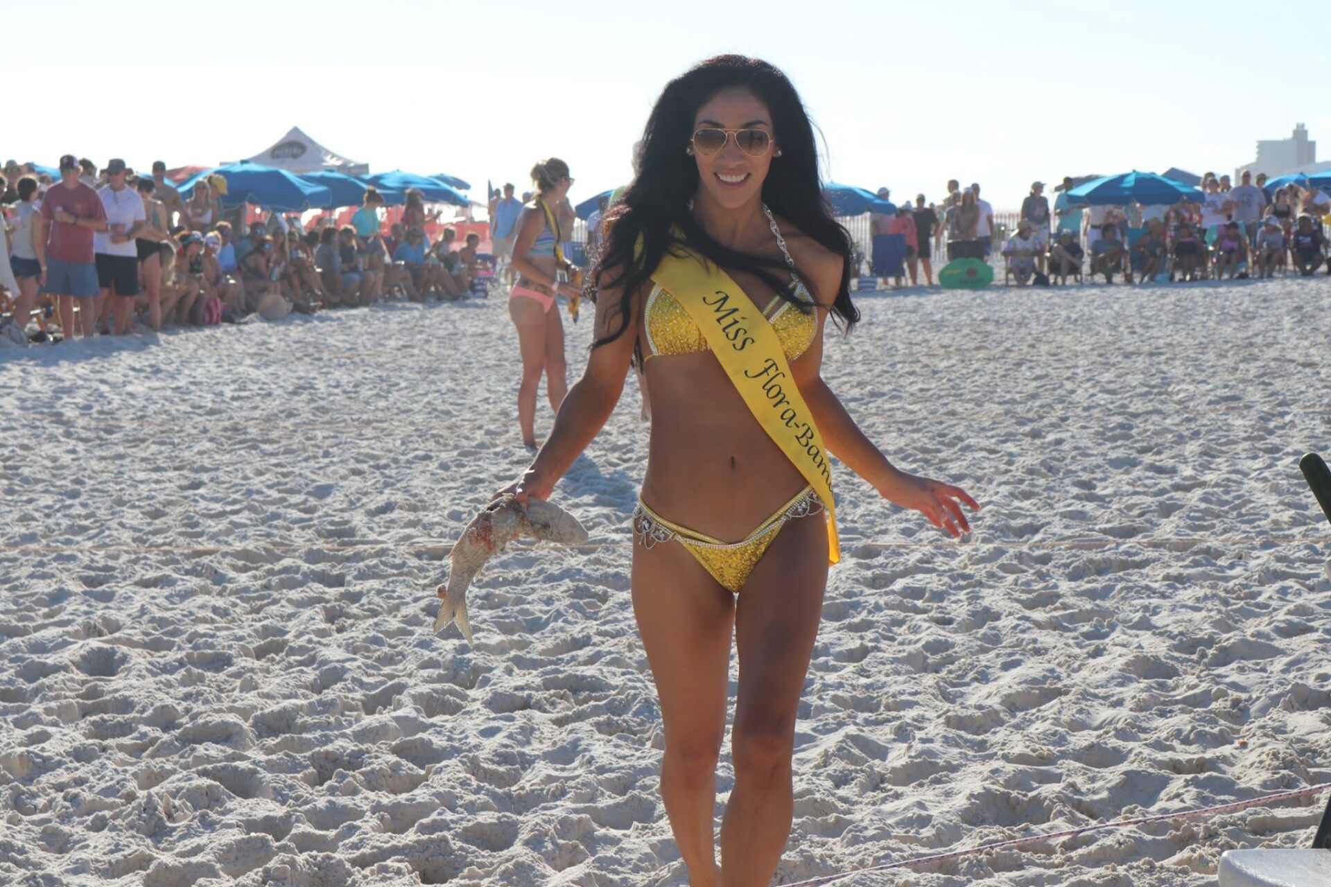 A contestant wearing a yellow sash and bikini walks across a white sand beach during an outdoor event.