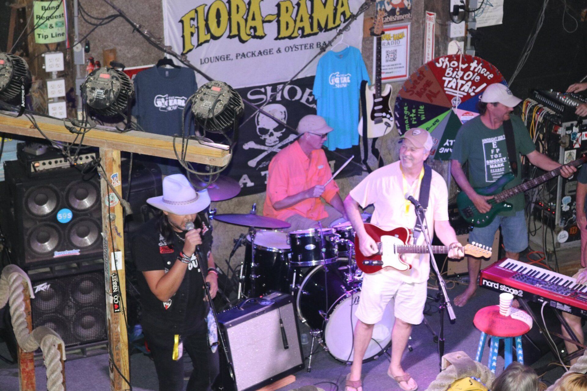 A band performs on a stage decorated with a Flora-Bama banner, featuring a singer, drummer, guitarist, and bassist.