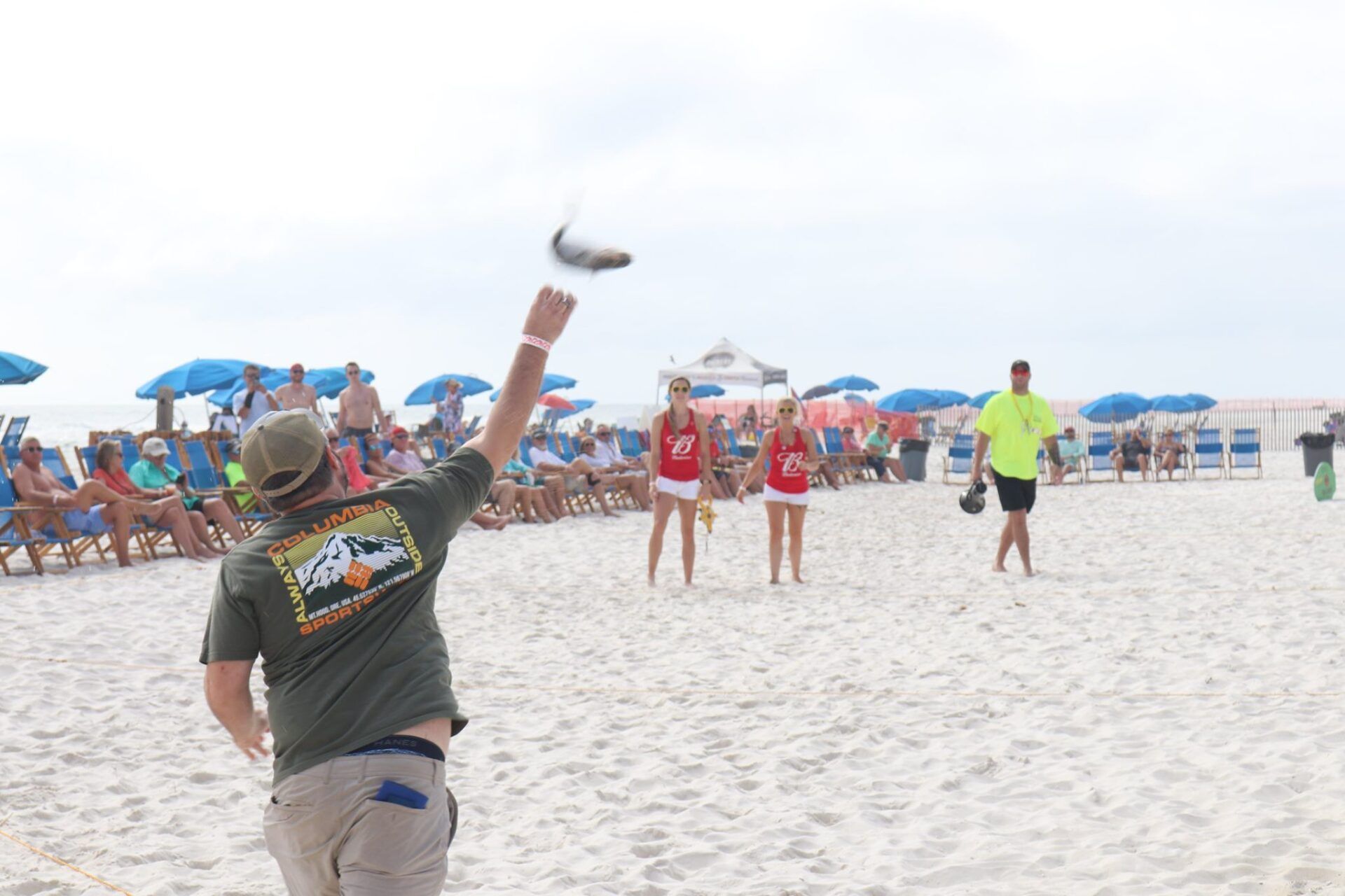 A person throws a fish into the air on a sandy beach crowded with spectators and blue umbrellas.
