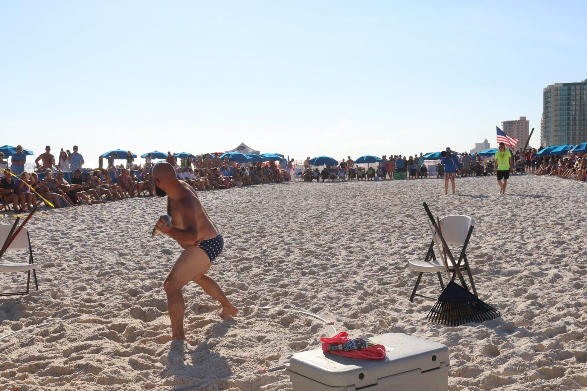 A person in swim trunks stands in a sand-covered area on a crowded beach with blue umbrellas and buildings in the distance.