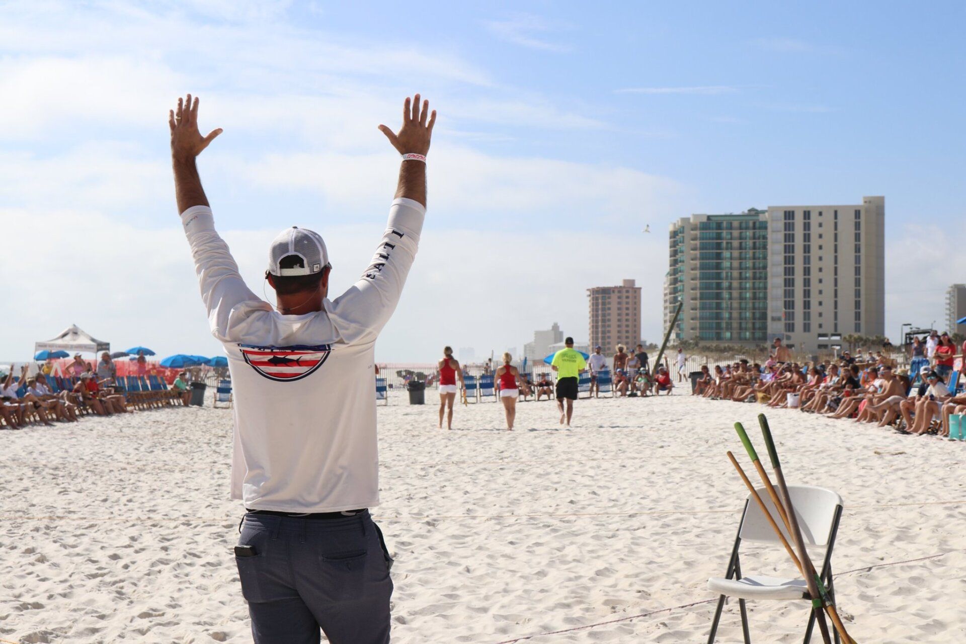 A person stands with arms raised on a beach, facing toward a small group of people in red uniforms and a referee.