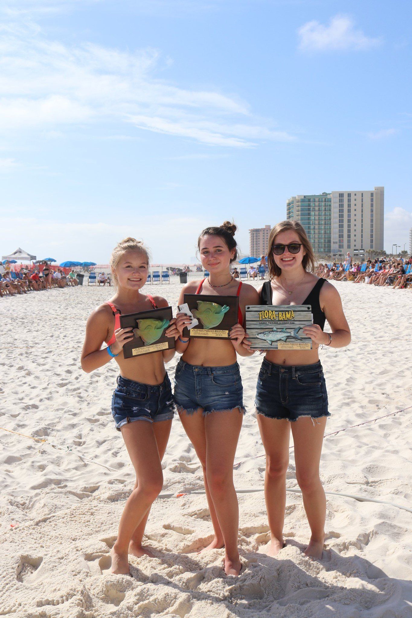 Three people stand on a sunny, crowded beach holding award plaques, wearing bikini tops and denim shorts.