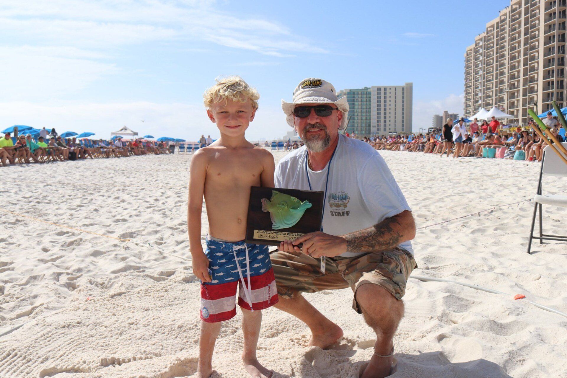A child and an adult hold a plaque together on a sandy beach near high-rise buildings.