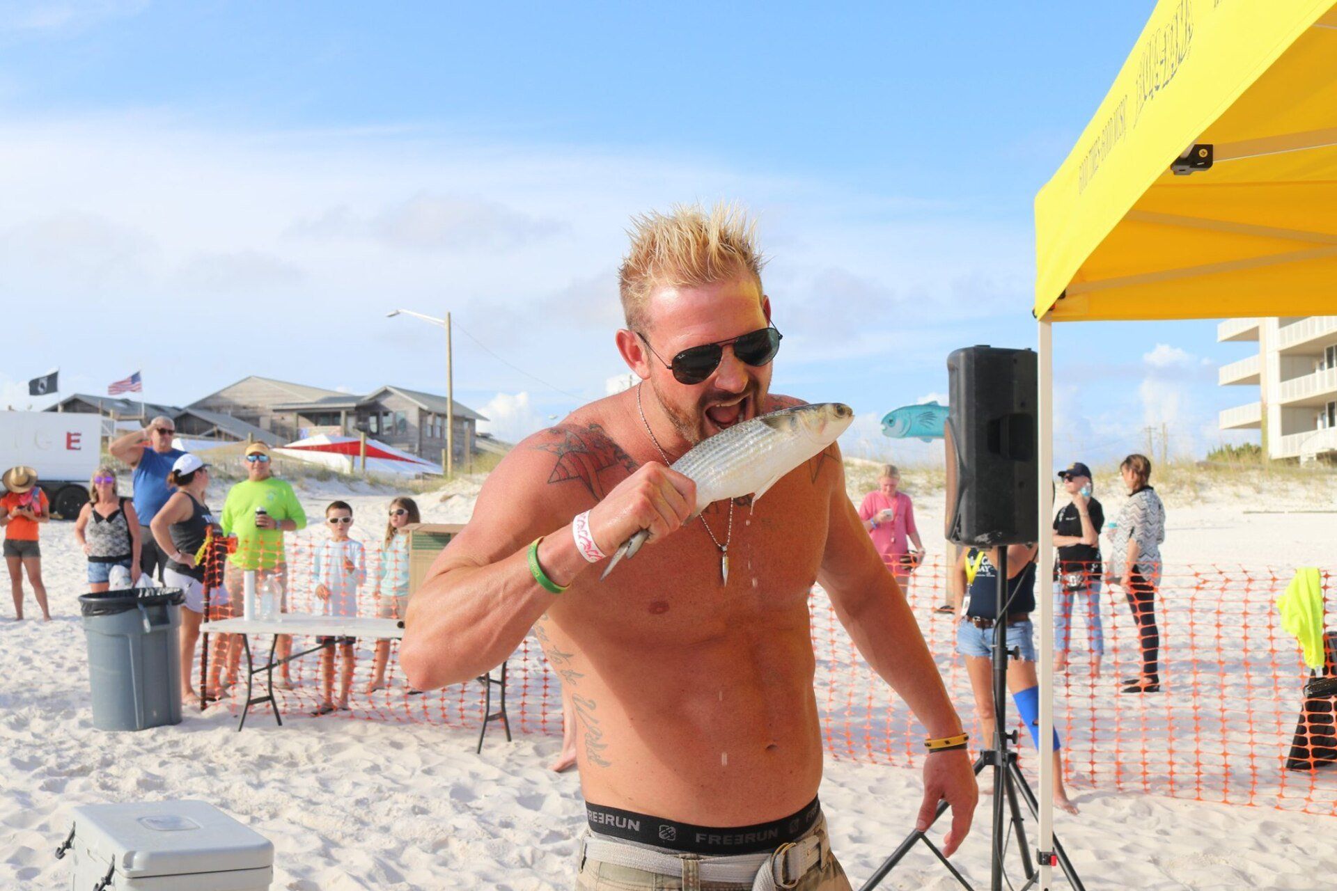 A shirtless person with sunglasses bites into a raw fish at a sunny beach festival under a yellow canopy.