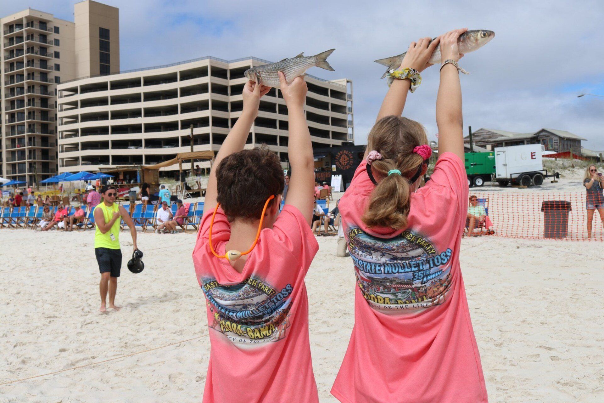 Two people in matching coral shirts stand on a sandy beach, holding fish high above their heads towards the sky.