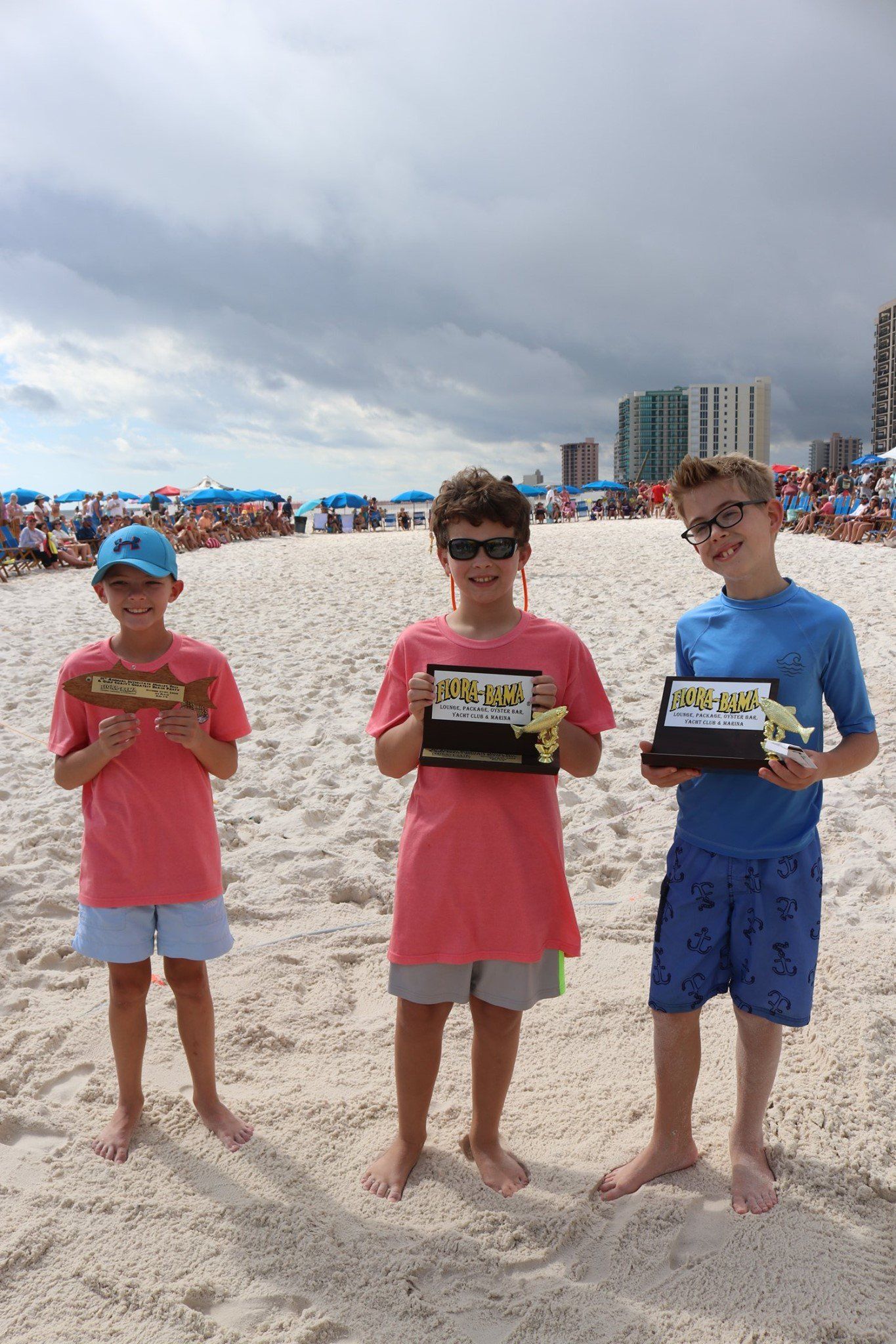 Three children holding awards while standing on a sandy beach under a cloudy sky.