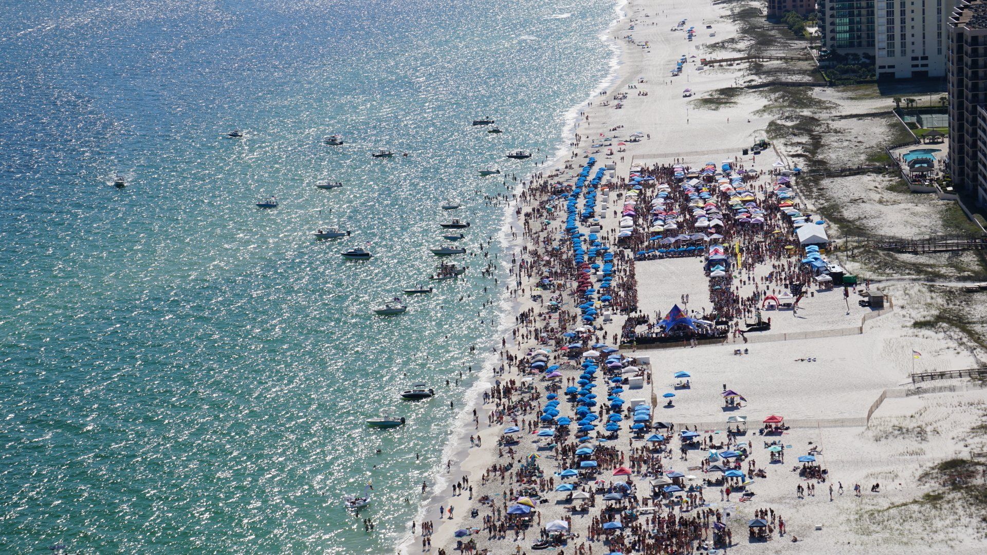 An aerial view of a crowded beach with many umbrellas, people, and several boats anchored in the nearby sparkling water.