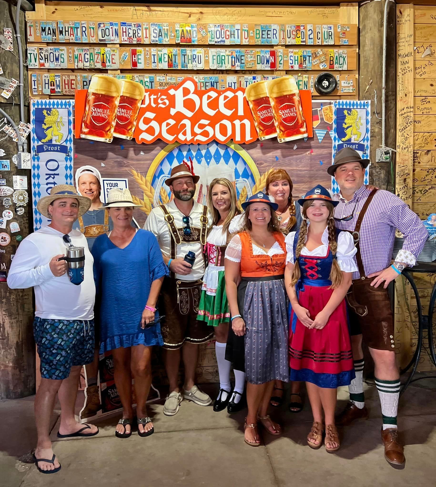 A group of people wearing traditional Oktoberfest attire and casual clothing pose in front of a 