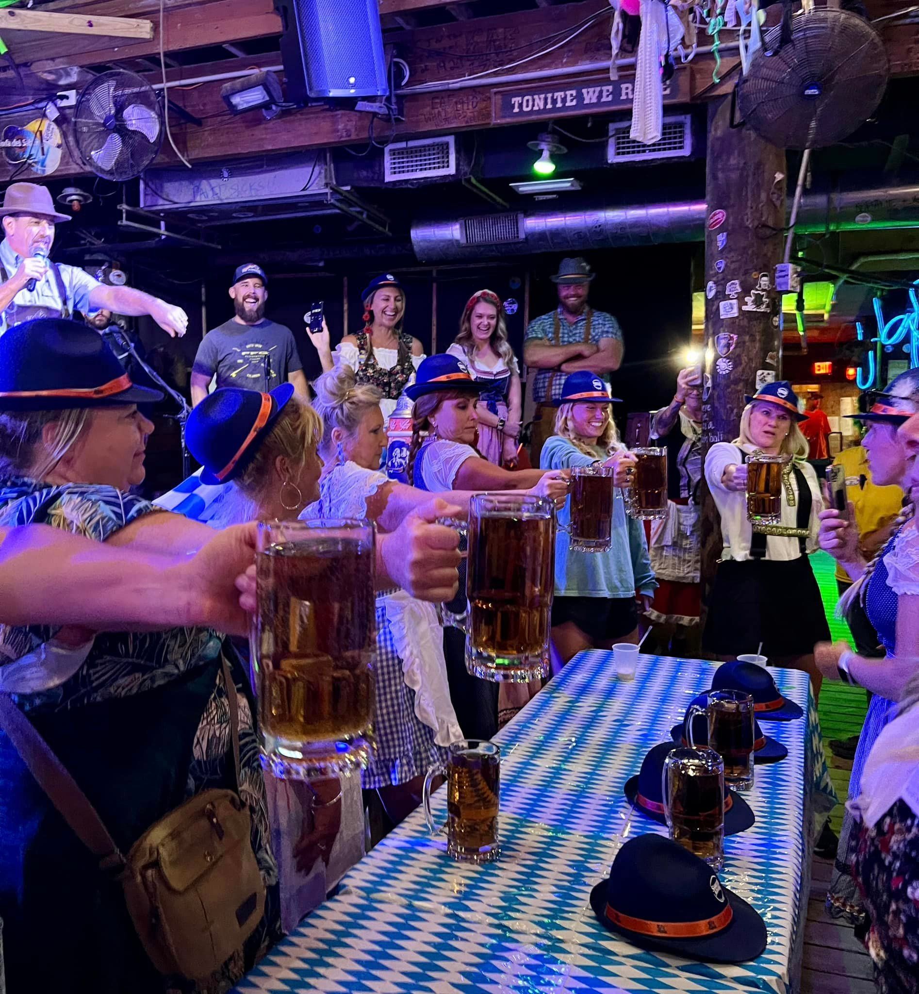 People in festive attire toast with beer steins at a table with a blue-and-white checkered tablecloth at an Oktoberfest.