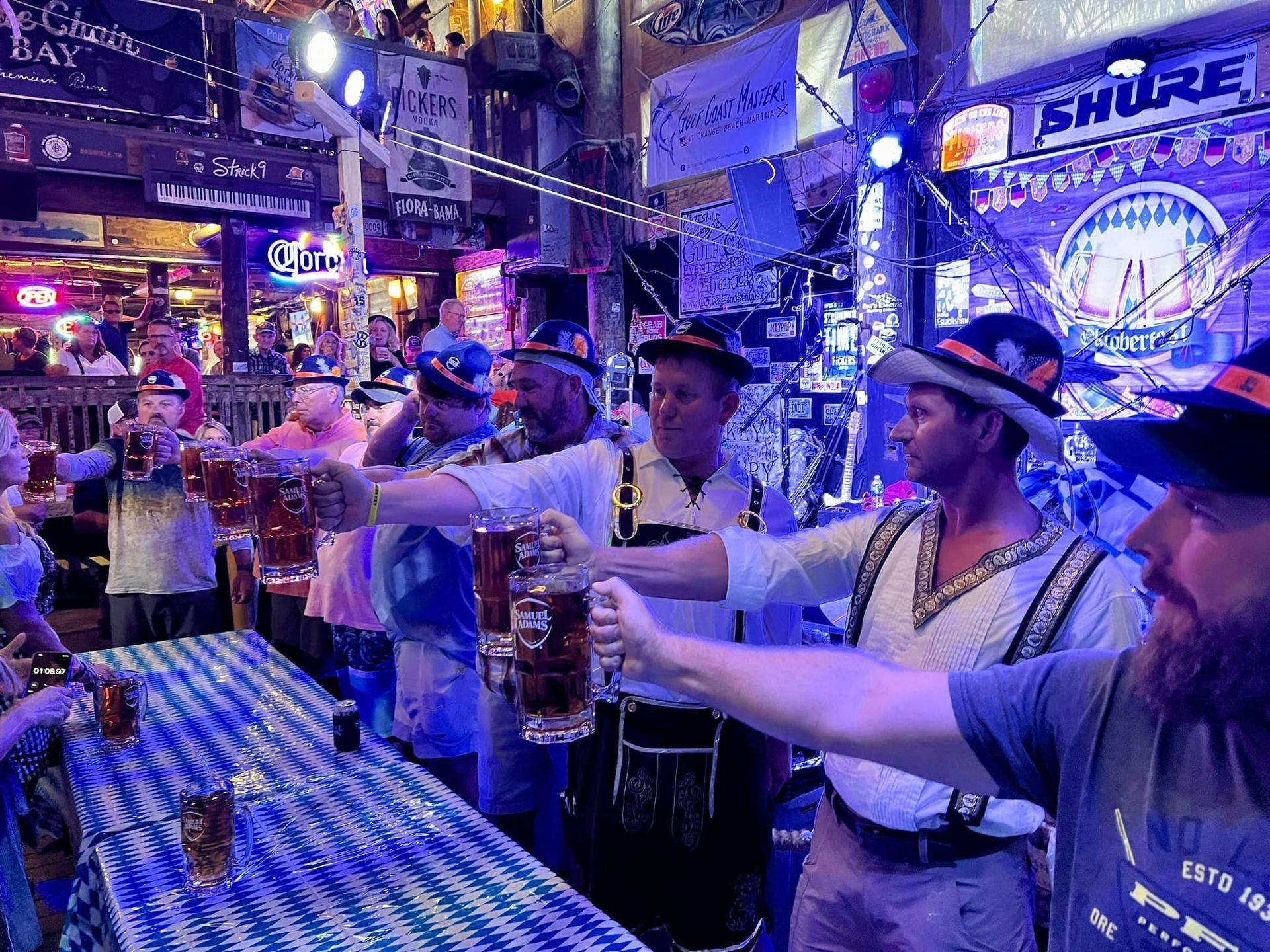 Group of people wearing hats and traditional attire toast with large beer steins at a table with a blue checkered cloth.