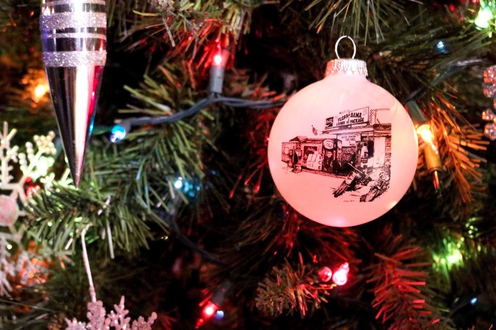 A round, white Christmas ornament with a dark building sketch hangs on a decorated tree with colorful lights.