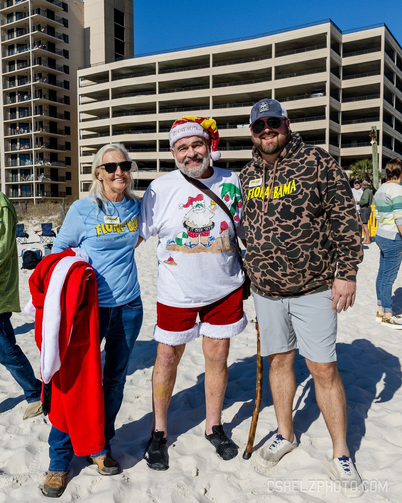 Three people stand on a sandy beach in front of a multi-story hotel, wearing sunglasses and holiday-themed casual attire.