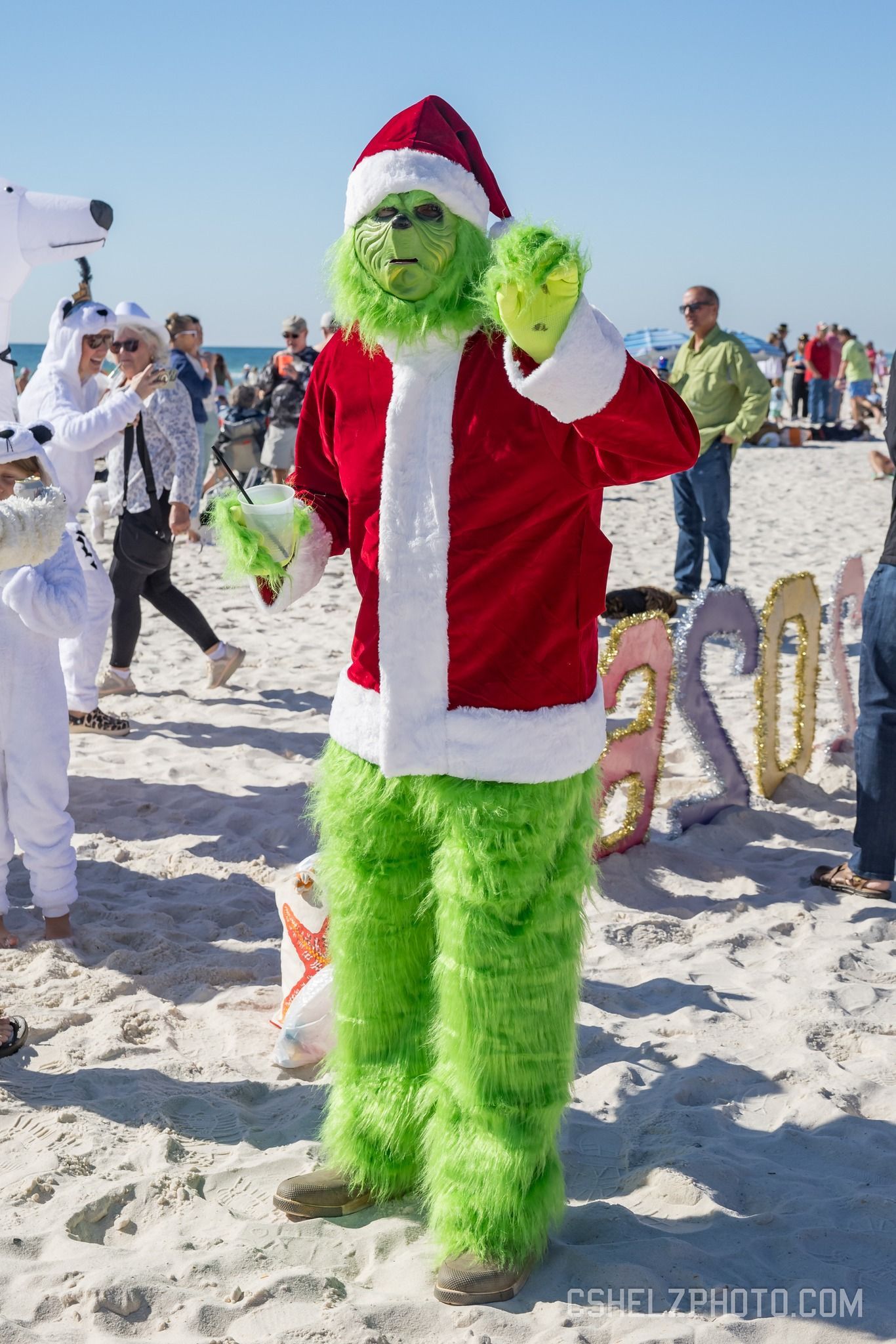 A person dressed in a green, fuzzy Grinch costume with a Santa hat and jacket standing on a sunny, sandy beach.