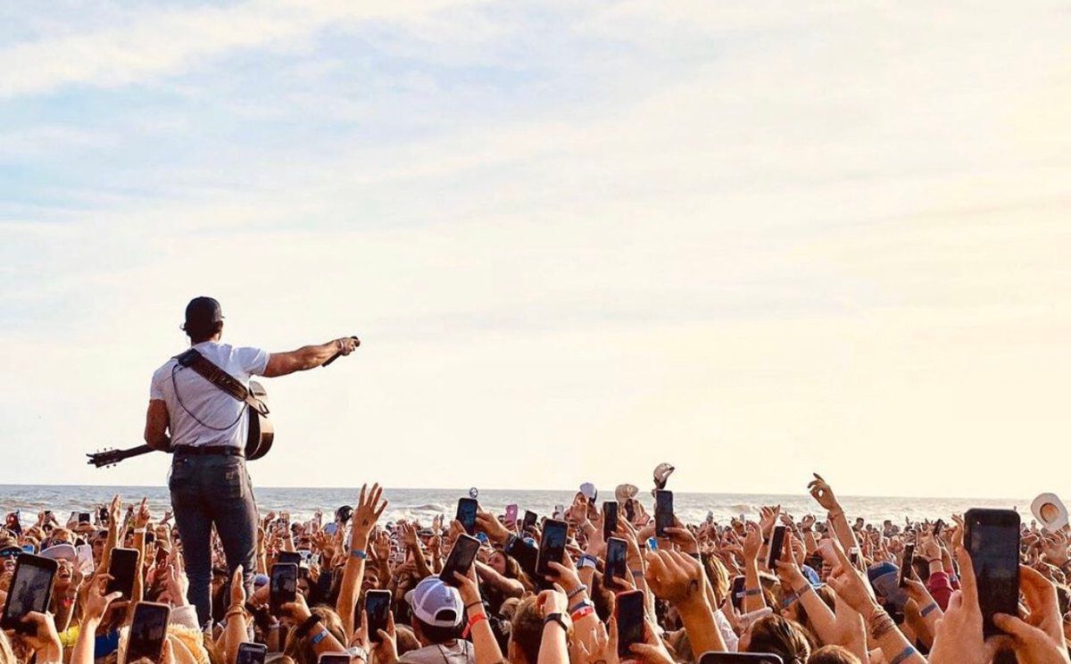 A musician stands on stage playing guitar for a large, cheering crowd at a beach concert under a bright, sunny sky.
