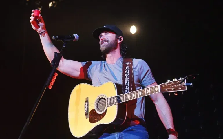 A musician in a cap and grey t-shirt performs on stage, holding a red cup aloft while playing an acoustic guitar.