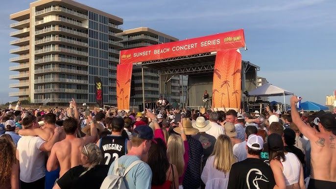 A crowd of people stands at a beach concert, facing a large, branded stage under a clear sky.