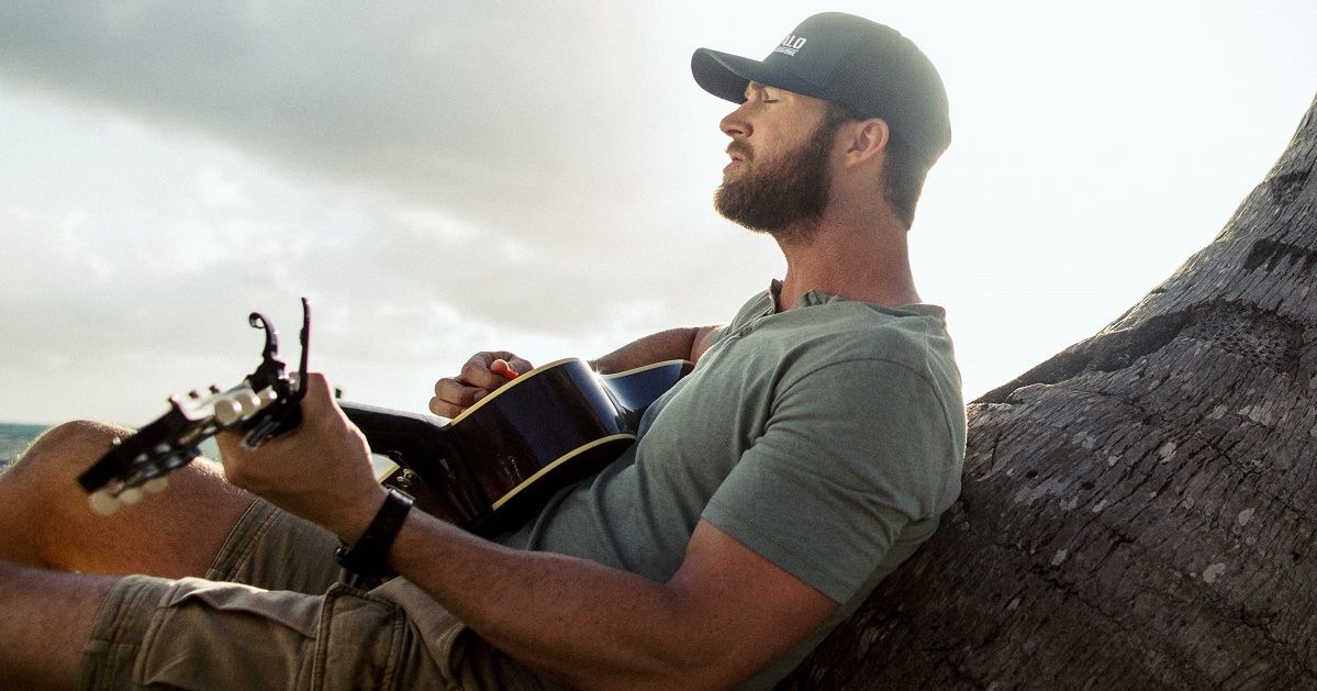 A person with a beard wearing a cap and olive t-shirt plays an acoustic guitar while sitting against a tree by the beach.