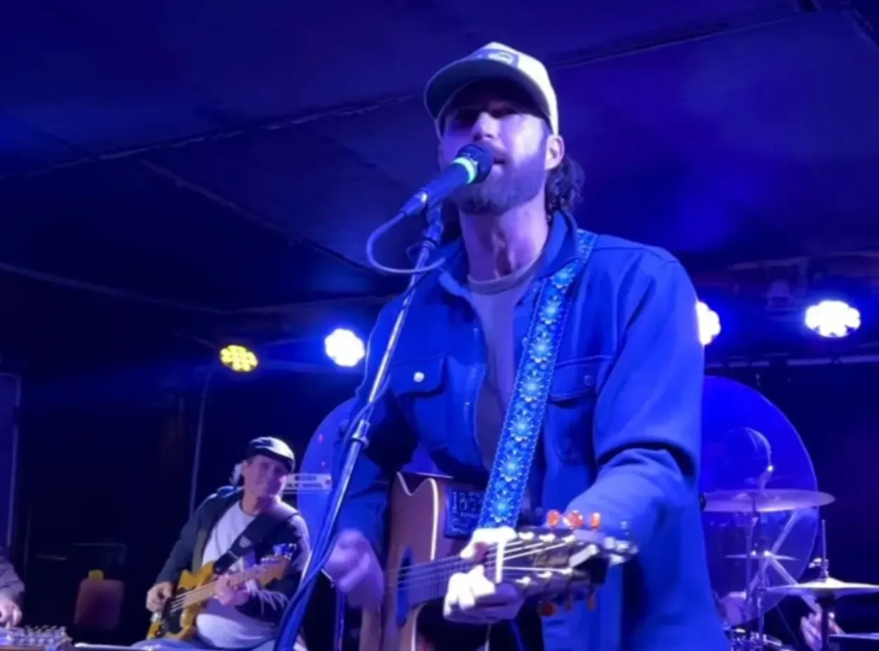 A musician plays acoustic guitar and sings into a microphone on a stage with blue lighting, with a bandmate behind him.