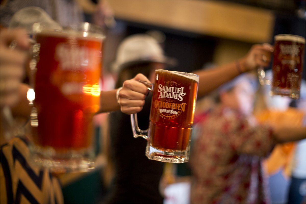 People raising Samuel Adams Oktoberfest beer mugs in a toast at a crowded indoor event.