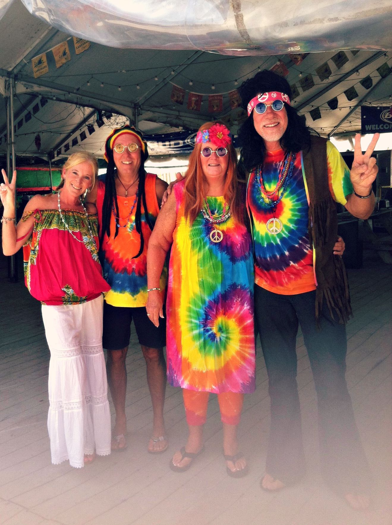 Four people posing in 1960s-style tie-dye outfits and hippie accessories under a tent at an outdoor event.