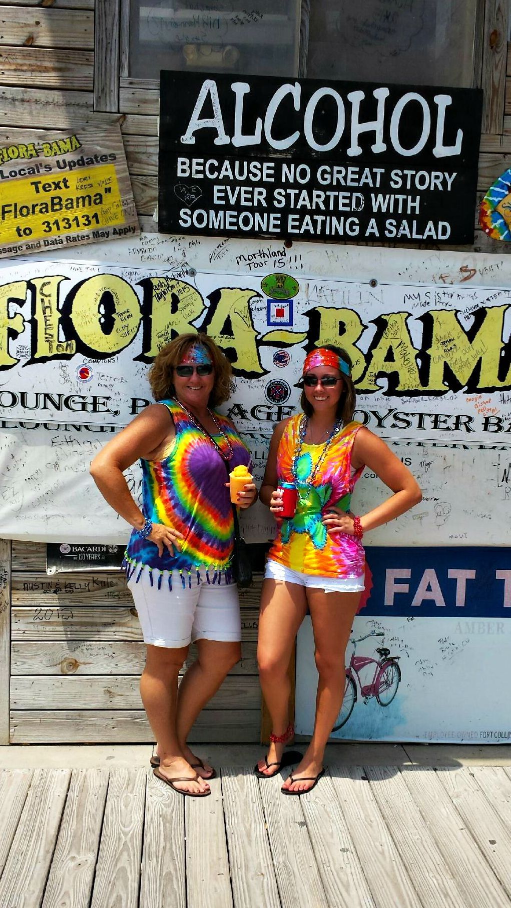 Two people in tie-dye shirts stand smiling in front of a wall featuring a sign that reads, 