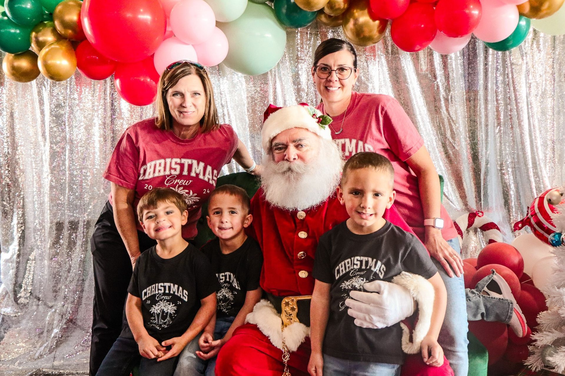 Santa Claus poses with two women and three children in matching Christmas shirts in front of a silver tinsel backdrop.