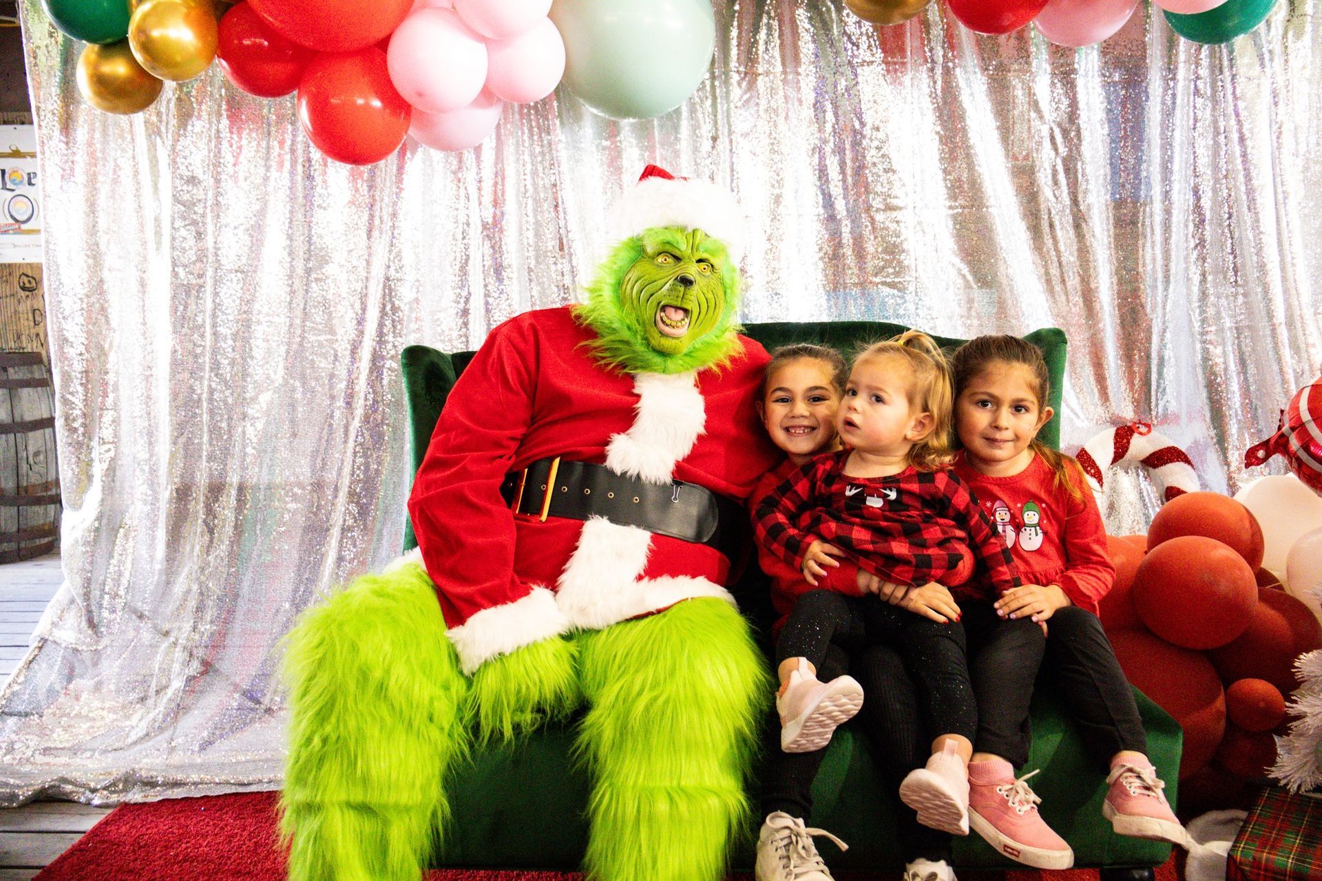 The Grinch in a Santa suit sits on a chair with three children before a silver curtain backdrop with festive balloons.