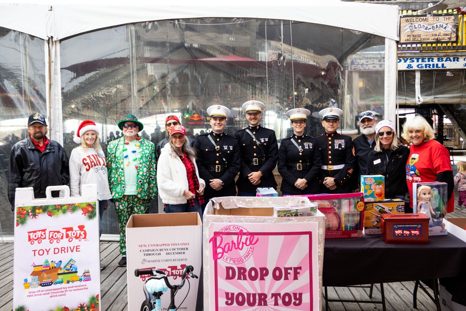 A group of people standing behind a toy drive donation station on a boardwalk, with Marine uniforms and holiday attire.