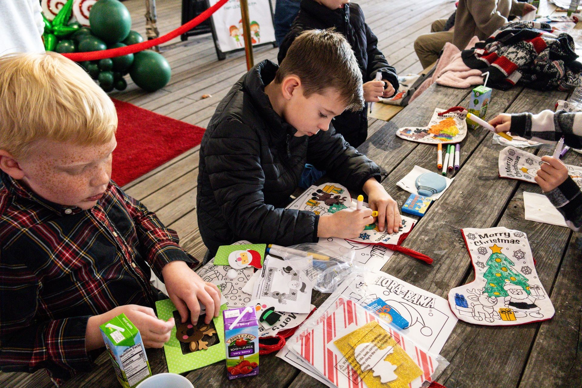 Children sit at a wooden picnic table coloring paper stockings and holiday-themed crafts in an outdoor setting.