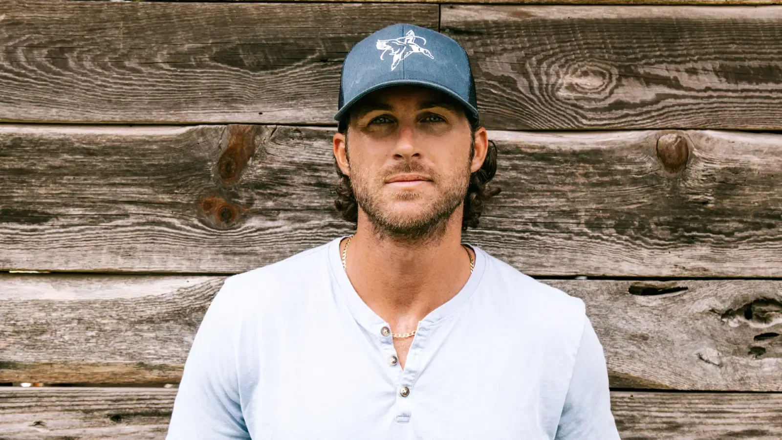 A person wearing a blue baseball cap and light-colored shirt stands in front of a rustic wooden wall.