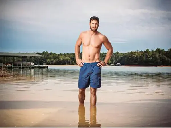 A shirtless person stands in shallow lake water, hands on hips, with a wooded shoreline in the background.