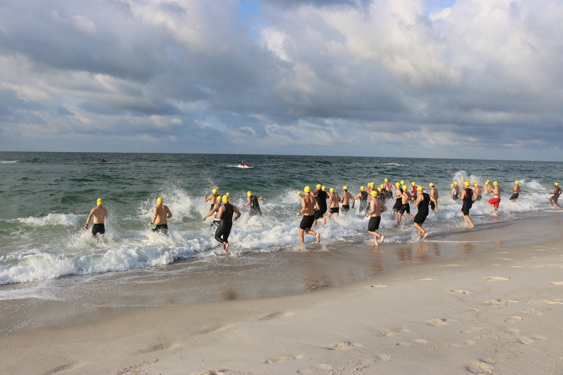 A group of swimmers wearing yellow caps run into the ocean waves on a sandy beach under a cloudy sky.