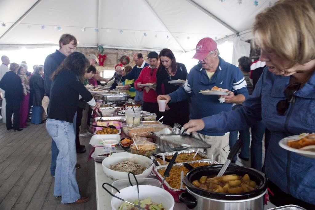 People in a tented pavilion serve themselves from a buffet line featuring various dishes in warming trays and bowls.